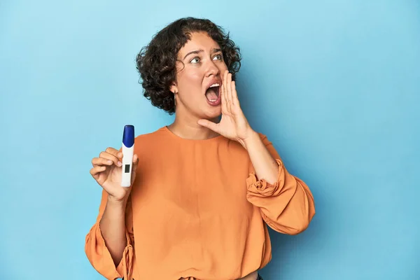 Young woman holding pregnancy test, studio background shouting and holding palm near opened mouth.