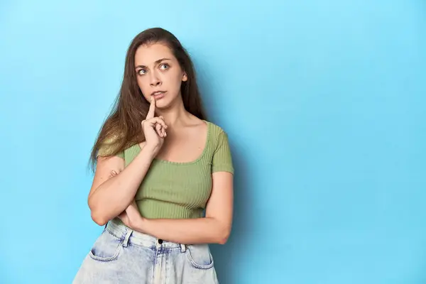 stock image Young Caucasian woman in a green top on a blue backdrop looking sideways with doubtful and skeptical expression.