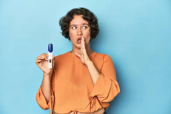 Young woman holding pregnancy test, studio background is saying a secret hot braking news and looking aside