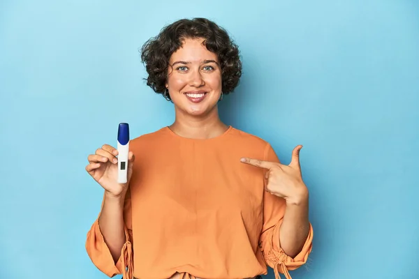 Young woman holding pregnancy test, studio background person pointing by hand to a shirt copy space, proud and confident
