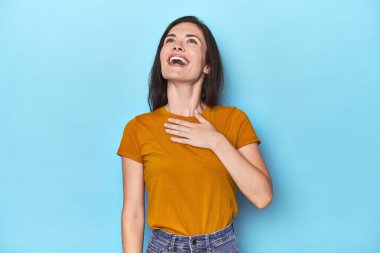 Mavi arka plandaki genç beyaz kadın kahkahalarla gülüyor elini göğsüne koyuyor.