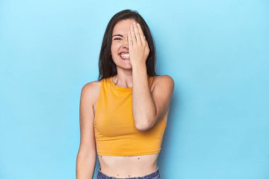 Mavi arka planda genç beyaz bir kadın yüzünün yarısını palmiyeyle örterek eğleniyor..