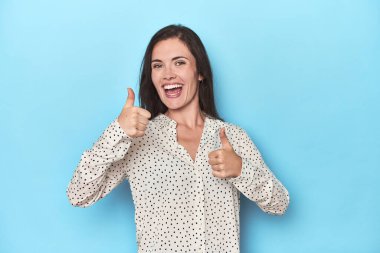 Mavi arka planda genç beyaz kadın iki parmağını da kaldırıyor, gülümsüyor ve kendinden emin..