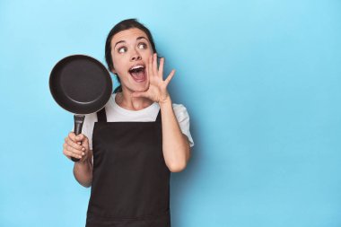 Mavi arka planda önlüğü ve tavası olan bir kadın bağırıyor ve avuç içini açık tutuyor..