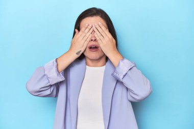 Mavi ceketli kadın mavi arka planda, elleri olan gözleri kapamaktan korkuyor..