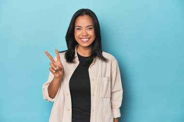 Mavi stüdyodaki Filipinli genç kadın zafer işareti gösteriyor ve gülümsüyor..