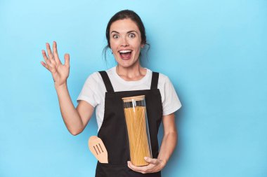 Mavi üzerinde makarna kavanozu olan aşçı kadın hoş bir sürpriz alıyor, heyecanlı ve el kaldırıyor..