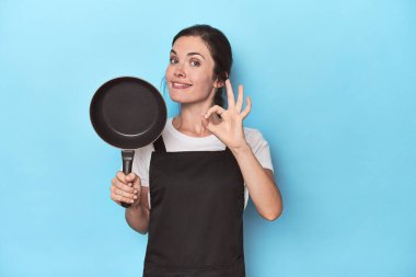 Mavi arka planda önlüğü ve tavası olan neşeli ve kendine güveni tam bir jest yapan bir kadın..