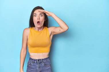 Mavi arka plandaki genç beyaz kadın yüksek sesle bağırıyor, gözlerini açık ve ellerini gergin tutuyor..