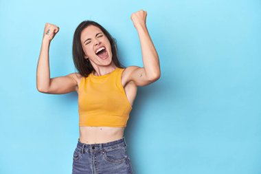 Mavi arka planda genç beyaz bir kadın zaferden sonra yumruğunu kaldırıyor, kazanan konsept..