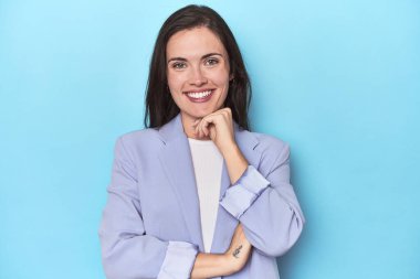 Mavi ceketli kadın mavi arka planda gülümsüyor mutlu ve kendinden emin, elleriyle çenesine dokunuyor..