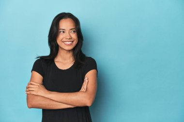 Mavi stüdyodaki Filipinli genç kadın kollarını kavuşturarak gülümsüyor..