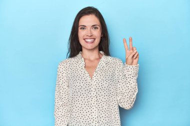 Mavi arka plandaki genç beyaz kadın neşeli ve tasasız parmaklarıyla barış sembolü gösteriyor..