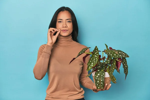 Filipina holding a plant on blue studio with fingers on lips keeping a secret.