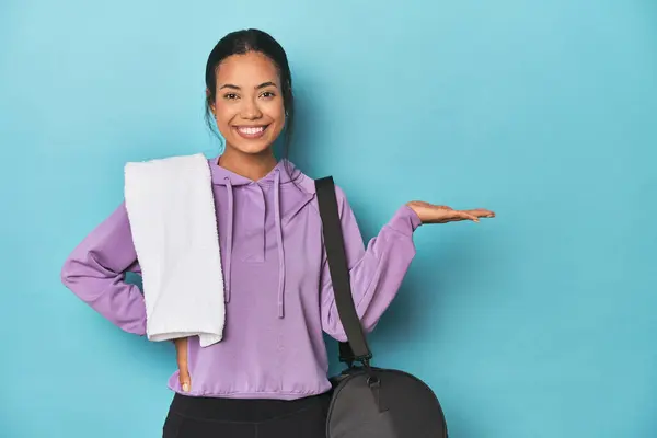 Filipina with gym gear on blue studio showing a copy space on a palm and holding another hand on waist.