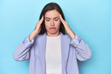 Mavi ceketli kadın mavi arka planda şakaklara dokunuyor ve başı ağrıyor..