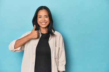 Mavi stüdyodaki Filipinli genç kadın gülümsüyor ve başparmağını kaldırıyor.