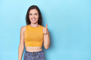 Mavi arka planda gülümseyen ve baş parmağını kaldıran beyaz bir kadın.
