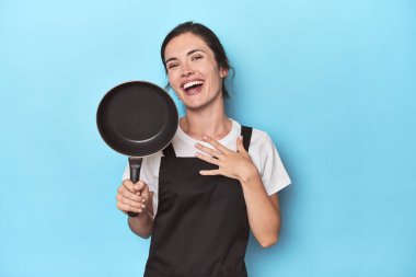 Mavi arka planda önlüğü ve tavası olan kadın kahkahalarla gülüyor elini göğsüne koyuyor..