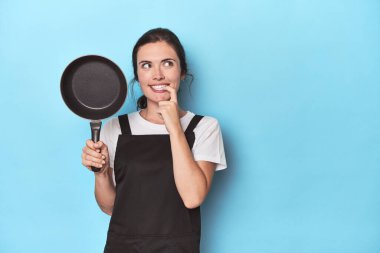 Mavi arka planda önlüğü ve tavası olan kadın rahat bir şekilde fotokopi odasına bakan bir şey düşünüyor..