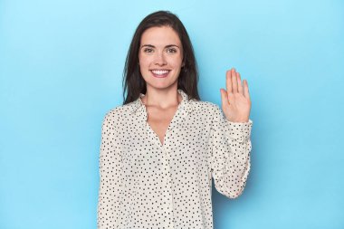 Mavi arka plandaki genç beyaz kadın gülümseyerek beş numarayı parmaklarıyla gösteriyor..