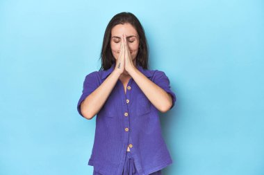 Mavi arka planda el ele tutuşup dua eden genç beyaz kadın, kendine güveniyor..