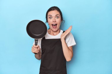 Mavi arka planda önlüğü ve tavası olan kadın şaşırdı ve şok oldu..