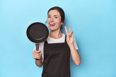 Mavi arka planda önlüğü ve tavası olan kadın iki numarayı parmaklarıyla gösteriyor..