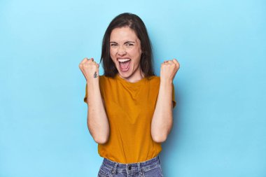 Mavi arka plandaki genç beyaz kadın tasasız ve heyecanlı tezahürat yapıyor. Zafer konsepti.