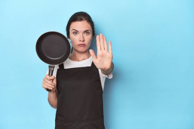 Mavi arka planda önlüğü ve tavası olan bir kadın elini uzatmış, dur işareti göstererek seni engelliyor..