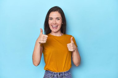 Mavi arka planda genç beyaz kadın iki parmağını da kaldırıyor, gülümsüyor ve kendinden emin..