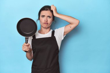 Mavi arka planda önlüğü ve tavası olan kadın şoka uğruyor. Önemli bir toplantıyı hatırlıyor..