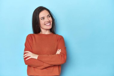 Mavi arka plandaki genç beyaz kadın kollarını kavuşturarak gülümsüyor..