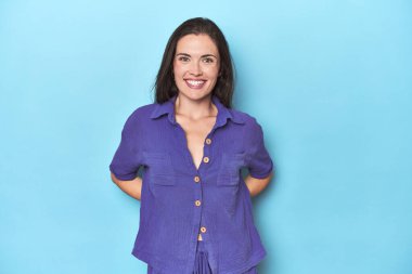 Mavi arka plandaki genç beyaz kadın mutlu, güler yüzlü ve neşeli.