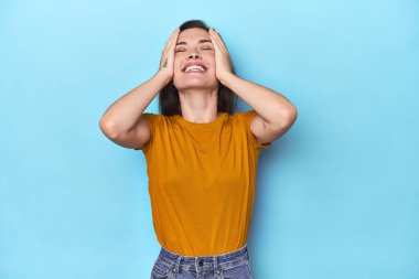 Mavi arka plandaki genç beyaz kadın mutlu bir şekilde ellerini başının üstünde tutarak gülüyor. Mutluluk kavramı.