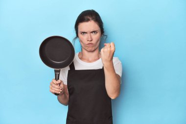 Mavi arka planda önlüğü ve tavası olan kadın kameraya yumruğunu gösteriyor, agresif yüz ifadesi var..