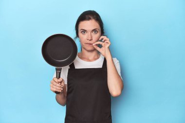 Önlüğü ve tavası mavi arka planda parmakları dudaklarında bir sır saklayan bir kadın..