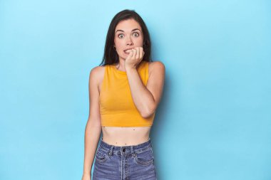 Mavi arka plandaki genç beyaz kadın tırnaklarını yiyor, gergin ve çok endişeli..