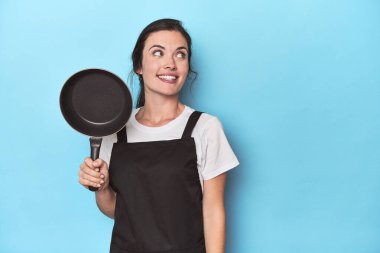 Mavi arka planda önlüğü ve tavası olan bir kadın gülümseyen, neşeli ve keyifli bir kenara bakar..