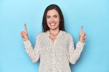 Mavi arka plandaki genç beyaz kadın işaret ediyor. İki parmağı da boş bir alan gösteriyor..