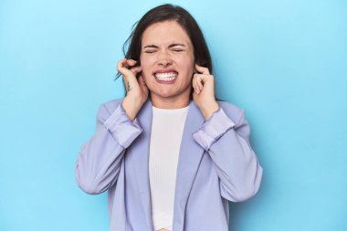 Mavi ceketli kadın mavi arka planda kulaklarını elleriyle kapatıyor..