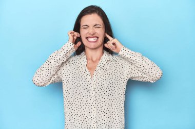 Mavi arka plandaki genç beyaz kadın kulaklarını elleriyle kapatıyor..