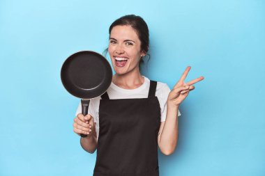 Mavi arka planda önlüğü ve tavası olan neşeli ve tasasız bir kadın parmakları olan bir barış sembolü gösteriyor..