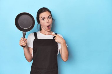 Mavi arka planda önlüğü ve tavası olan kadın yan tarafı işaret ediyor.