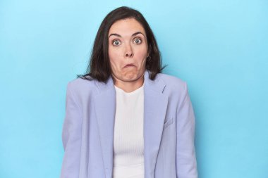 Mavi ceketli kadın mavi arka planda omuzlarını silker ve gözleri açık kalır..