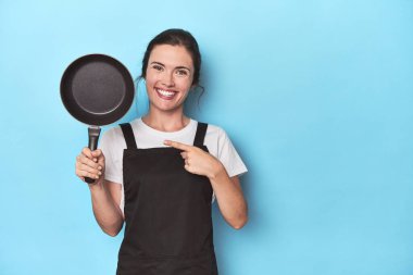 Mavi arka planda önlüğü ve tavası olan bir kadın gülümsüyor ve boş alanda bir şey gösteriyor..