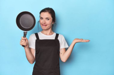 Mavi arka planda önlüğü ve tavası olan bir kadın avucunun içinde bir fotokopi alanı gösteriyor ve belinde başka bir el tutuyor..