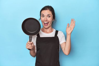 Mavi arka planda önlüğü ve tavası olan kadın hoş bir sürprizle karşılaşıyor, heyecanlı ve el kaldırıyor..