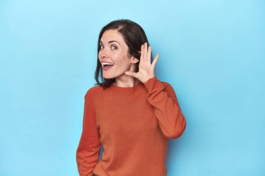 Mavi arka planda genç beyaz kadın bir dedikoduyu dinlemeye çalışıyor..