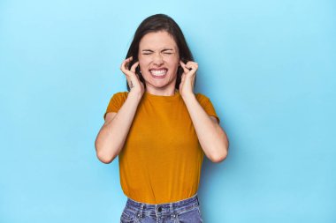 Mavi arka plandaki genç beyaz kadın kulaklarını elleriyle kapatıyor..